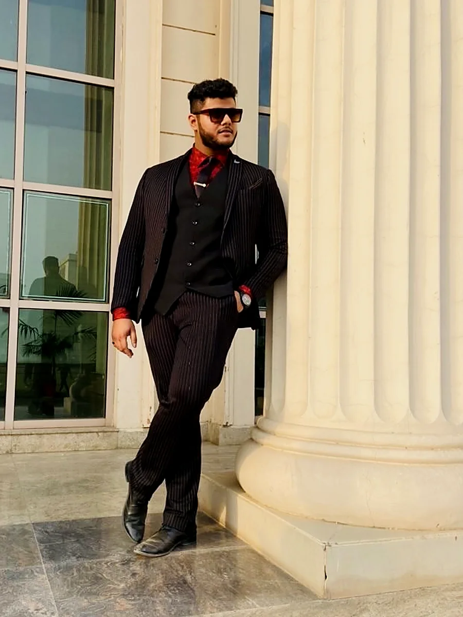 Amritash Chowdhry in a black pinstripe three-piece suit and red shirt, leaning on a stone column