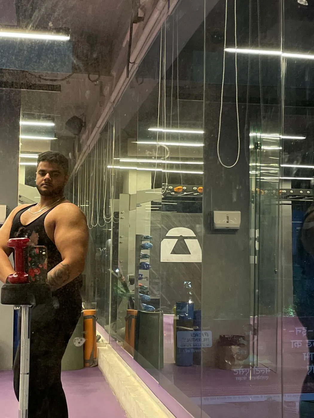 Amritash in a black tank top flexing in a gym mirror, side view