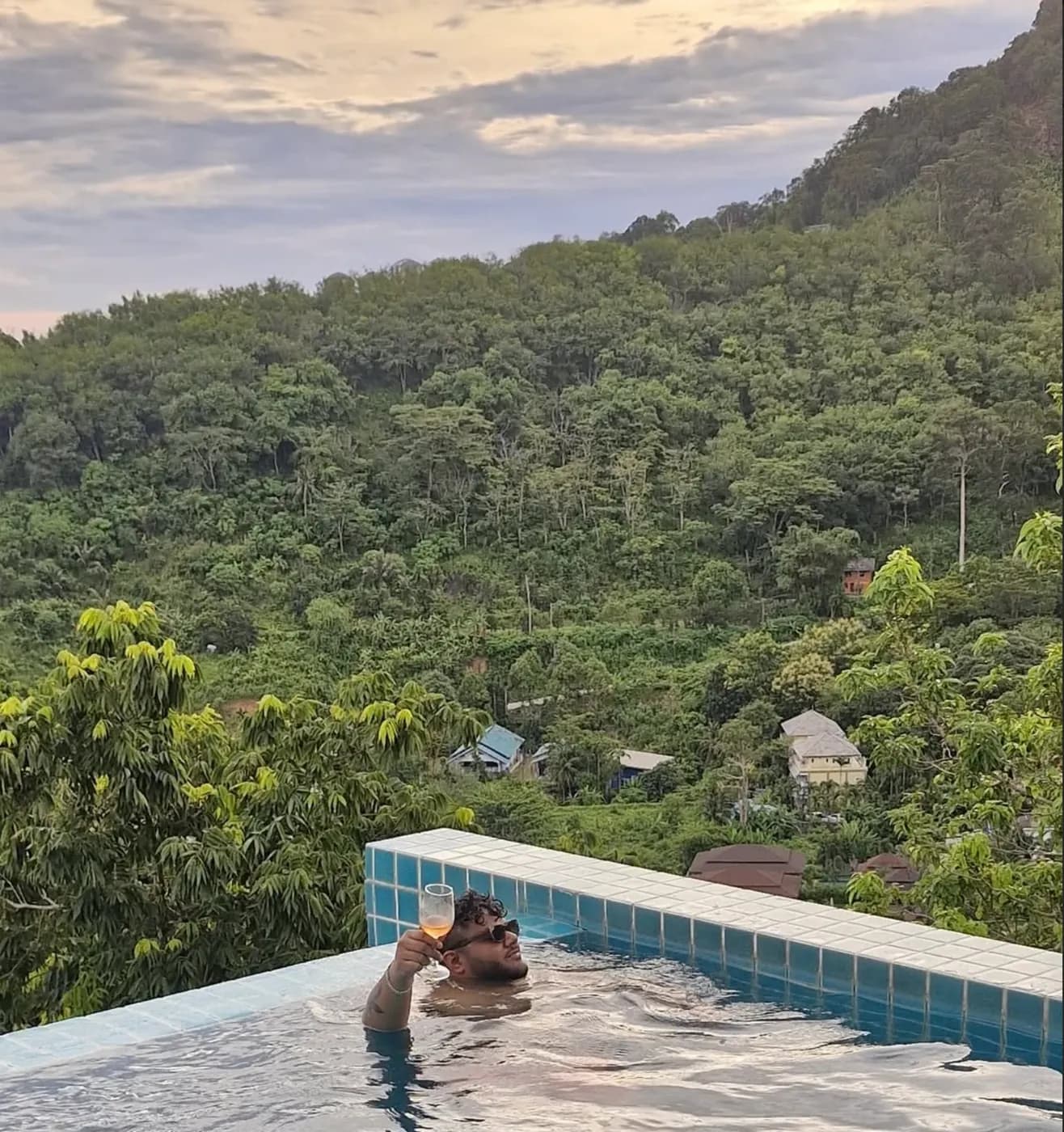 Amritash in an infinity pool raising a glass, green hills behind