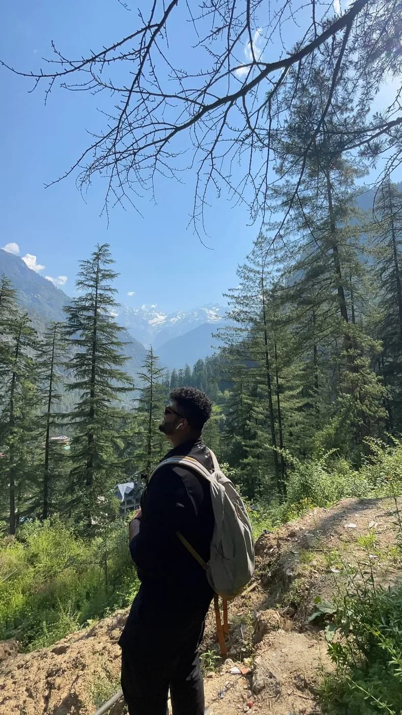 Amritash with a backpack among pine trees, looking toward snow-capped mountains