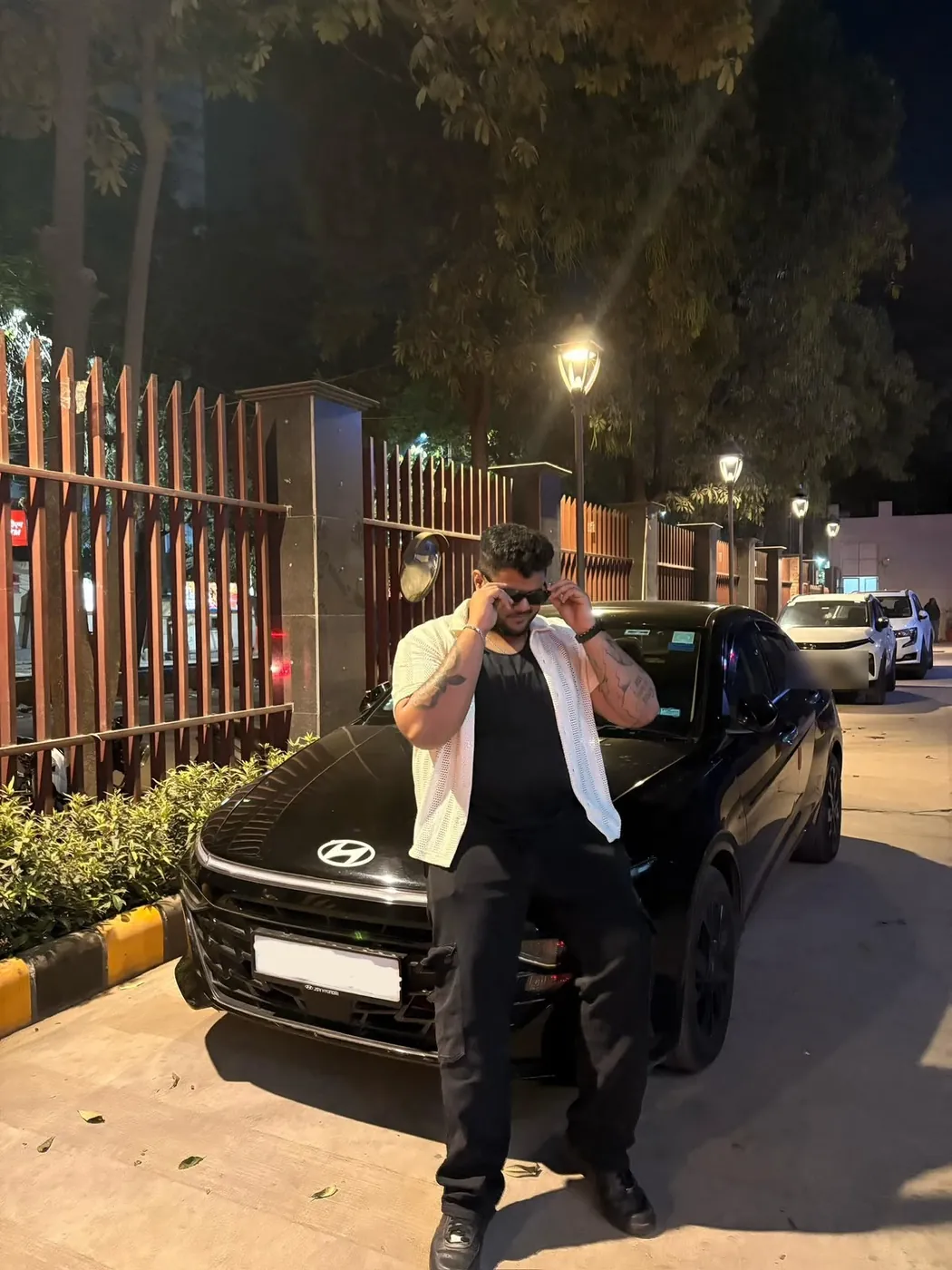 Amritash leaning on a black car at night, adjusting his sunglasses