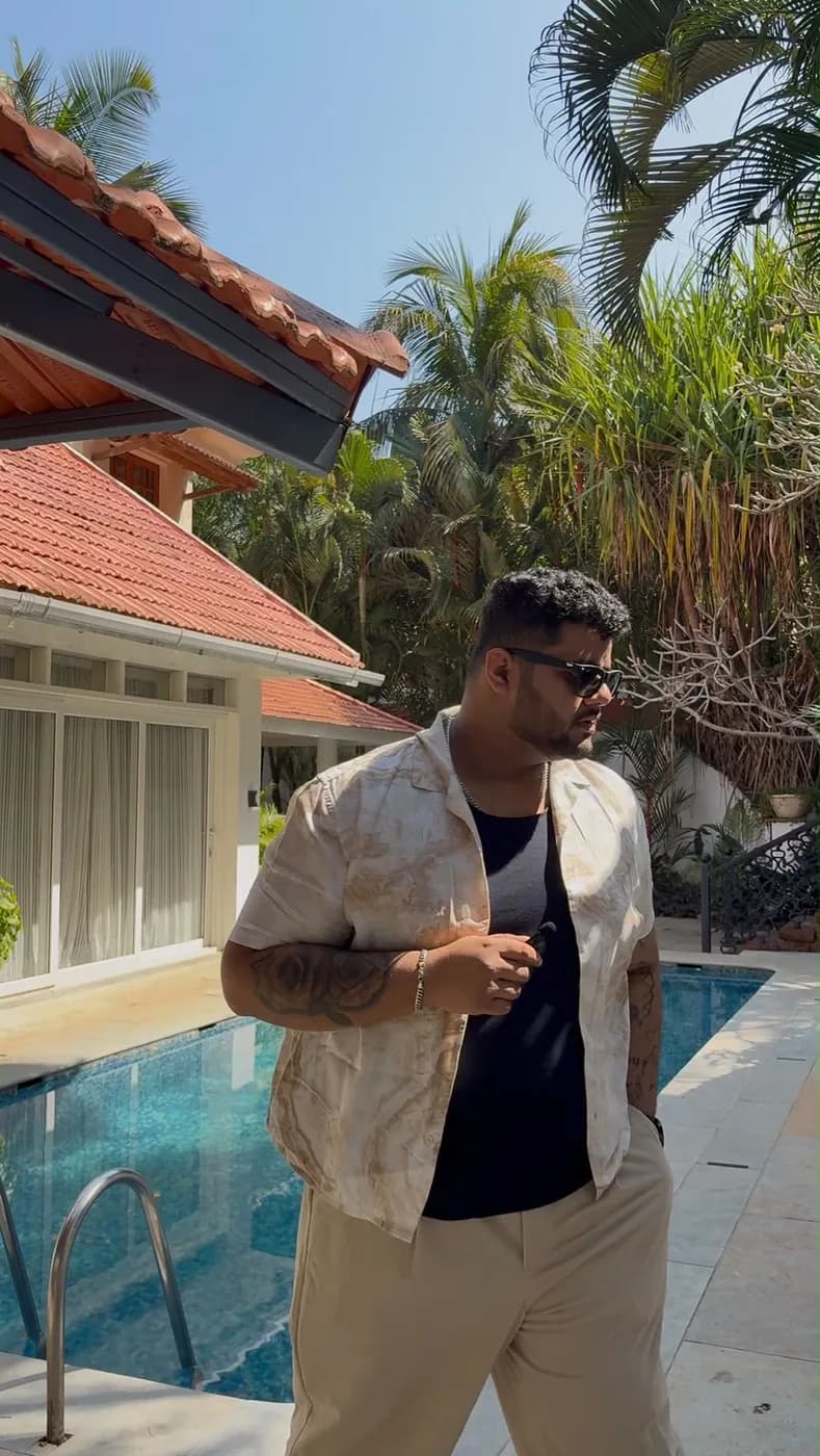 Amritash in a beige open shirt and black vest by a pool, looking to the side