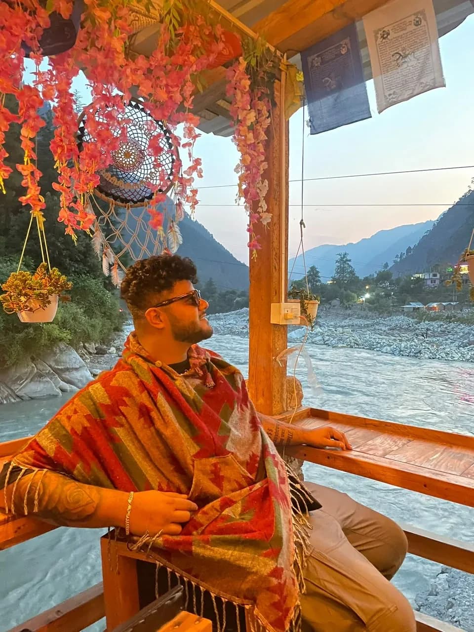 Amritash wrapped in a patterned shawl at a riverside café at dusk, mountains behind