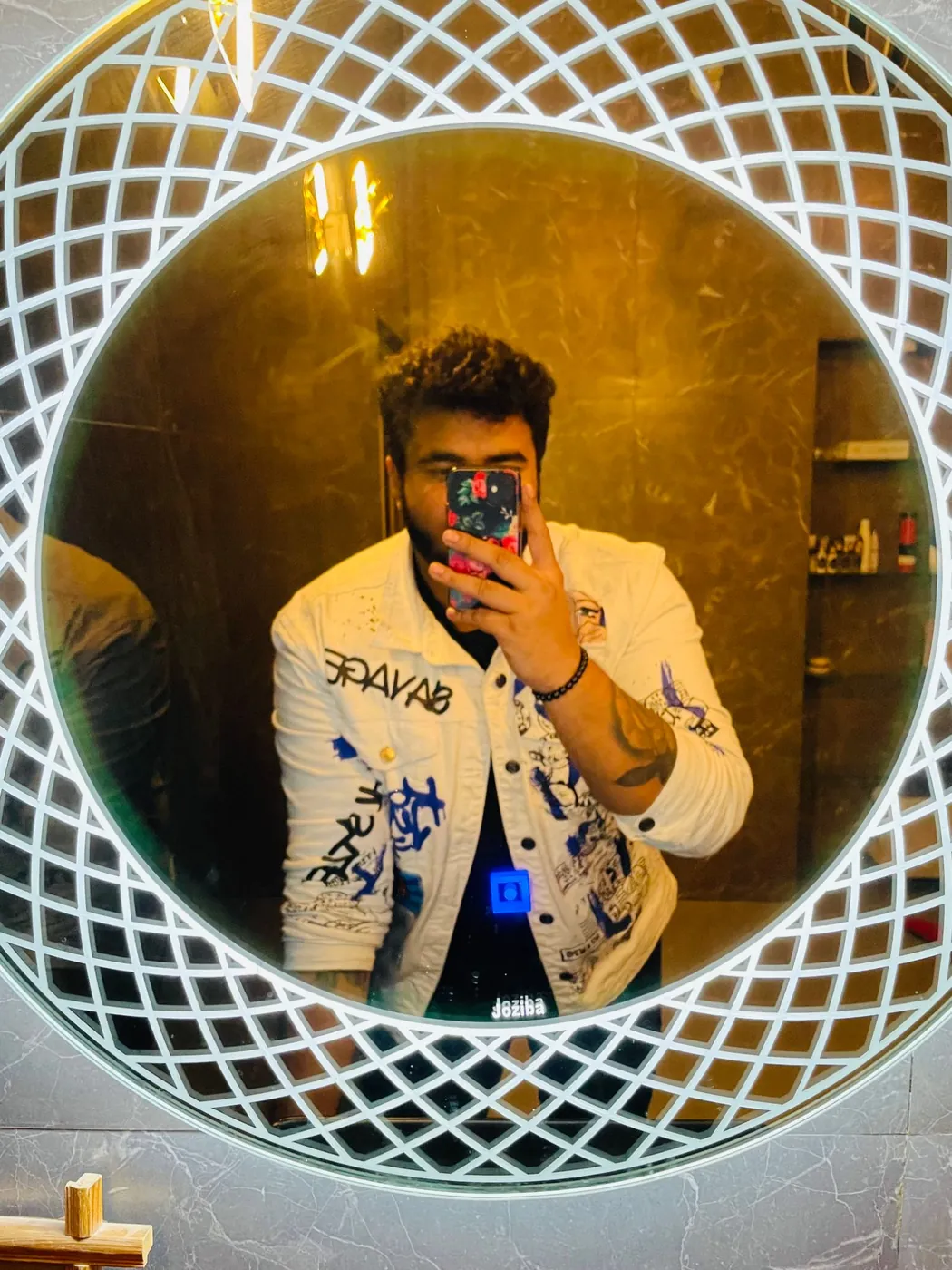 Amritash in a white graphic denim jacket, in a round lit mirror