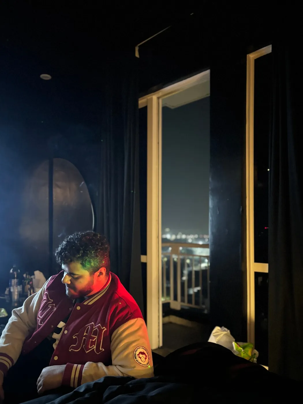 Amritash in a maroon varsity jacket in a dark room, city lights through the balcony door